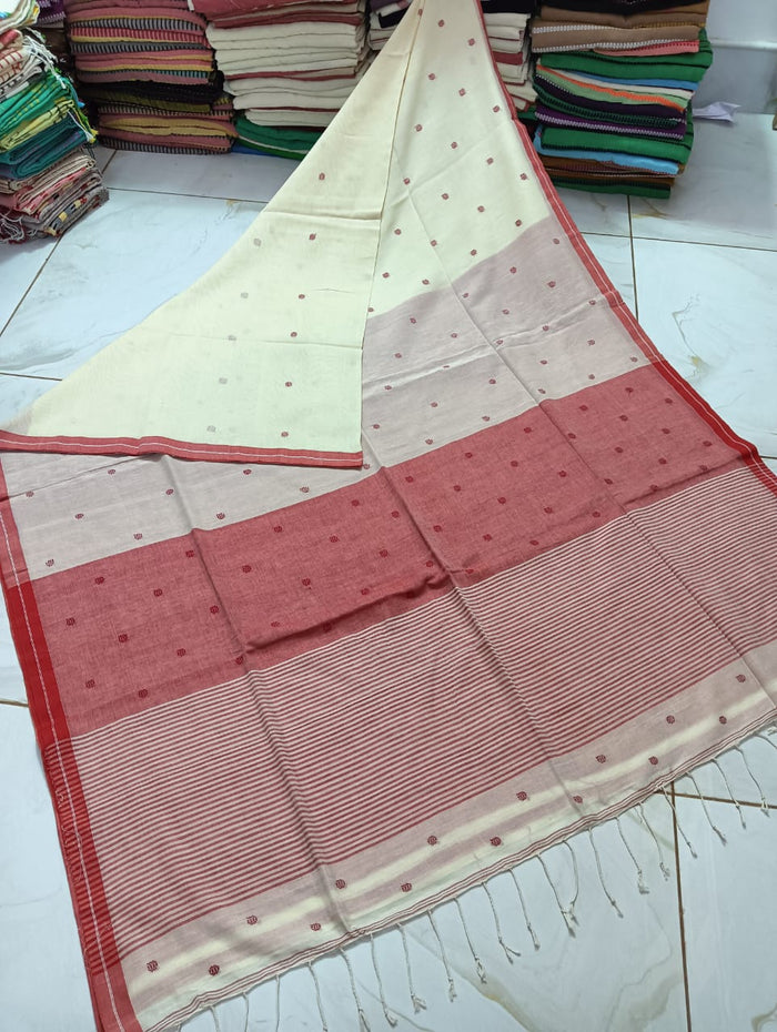 A folded white and red cotton jamdani saree with dobby weaving, displayed on a counter with various other sarees in the background.